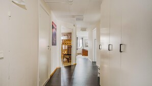 Un pasillo largo y estrecho con piso oscuro, paredes blancas y un techo con iluminación empotrada. Hay puertas a ambos lados y un banco de madera con cojines a lo lejos.