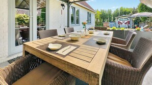 An outdoor dining area with a wooden table and wicker chairs, set against a white building with large windows. - Nice home in Bredene with heated swimming pool (Bredene)