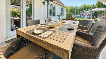 Un espacio de comedor al aire libre con una mesa de madera y sillas de mimbre, frente a un edificio blanco con grandes ventanales.