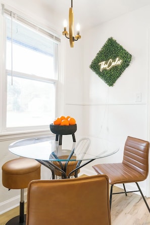 A dining area with a glass table, two brown leather chairs, a black bowl with oranges, and a chandelier.