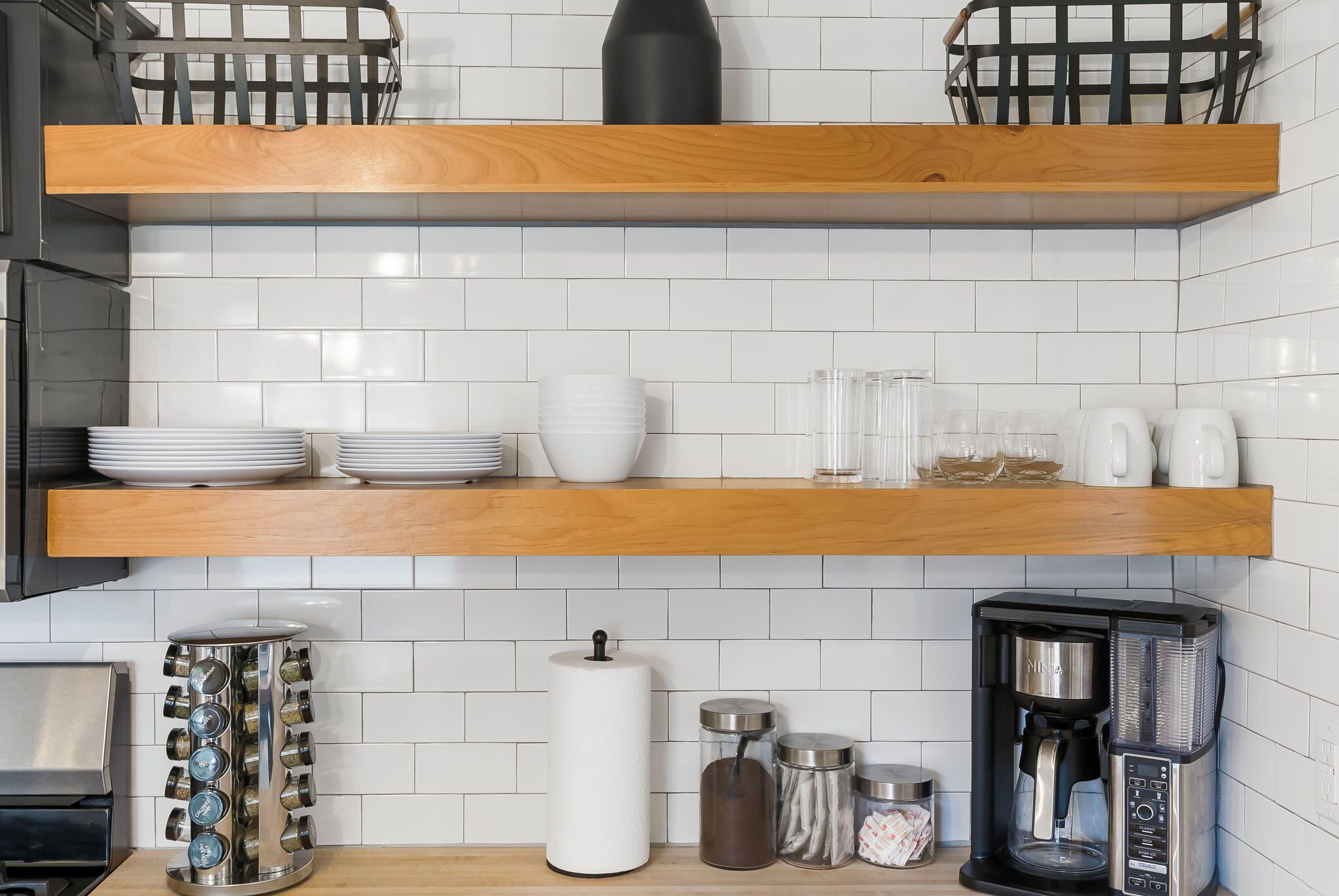 Une cuisine avec un dosseret de carreaux blancs, des étagères en bois et divers ustensiles de cuisine, notamment une machine à café et des récipients à épices.