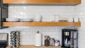 Une cuisine avec un dosseret de carreaux blancs, des étagères en bois et divers ustensiles de cuisine, notamment une machine à café et des récipients à épices.