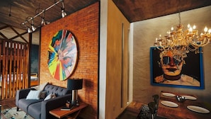 Una sala moderna con pared de ladrillo decorativa, un cuadro abstracto de colores, un sofá negro, una mesa de comedor con sillas y una araña.
