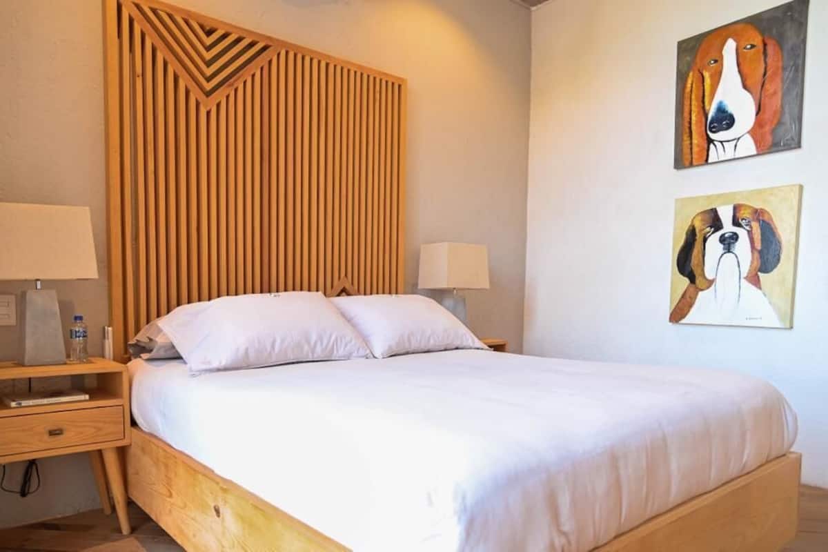 A bedroom with a wooden bed frame, two bedside tables with lamps, and two wall-mounted paintings of dogs.