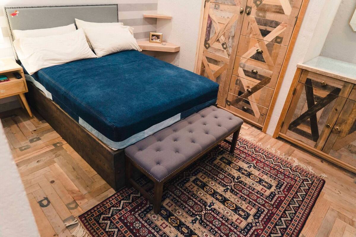 A bedroom with a bed, bench, wooden wardrobe, and patterned rug.