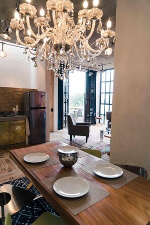 A dining area with a wooden table set for four, a chandelier above, and a kitchen with a refrigerator to the left. - Canela Fina Solo adultos (San Miguel de Allende)