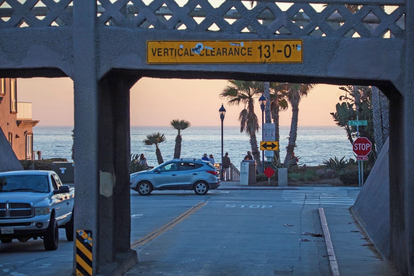 A coastal street with a vertical clearance sign, parked cars, and a view of the ocean.
