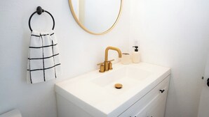 Un bagno con un lavandino bianco, un rubinetto dorato, un portasci con un asciugamano a quadretti, uno specchio rotondo e un dispenser per sapone.
