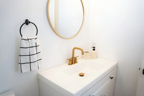 A bathroom with a white sink, gold faucet, a towel rack with a checkered towel, a round mirror, and a soap dispenser.