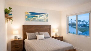 Una camera da letto con una testiera in legno, un letto con biancheria grigia, due comodini con lampade, un quadro appeso al muro e una finestra vista palme.