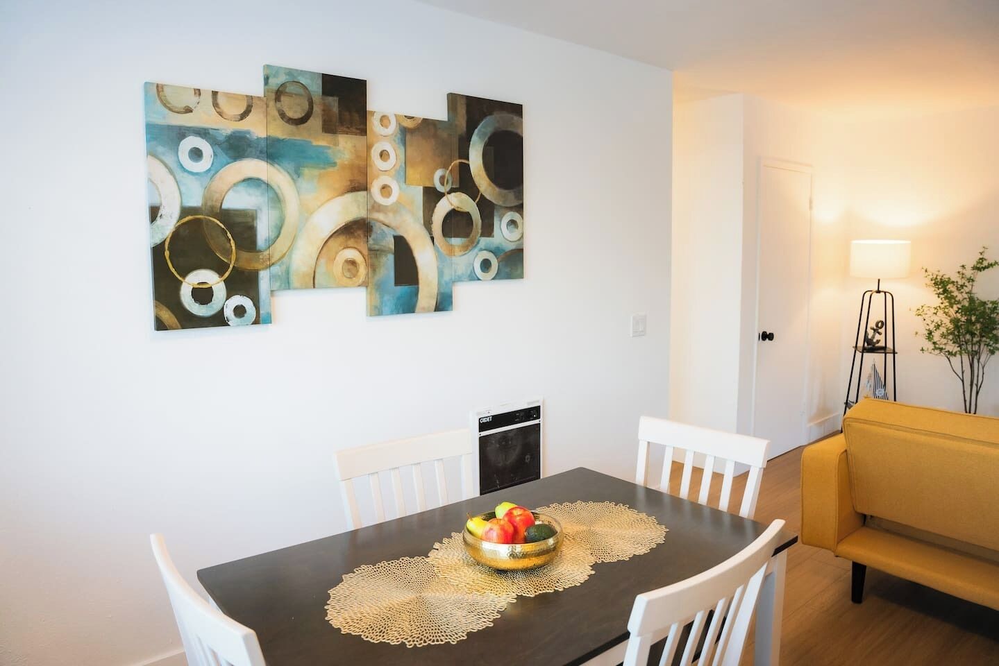 A dining area with a table set for two, a fruit bowl on the table, and a modern abstract painting on the wall.
