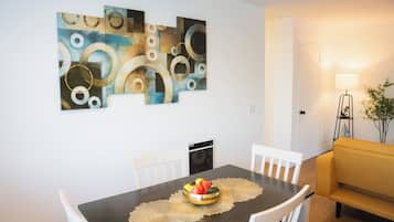 A dining area with a table set for two, a fruit bowl on the table, and a modern abstract painting on the wall.