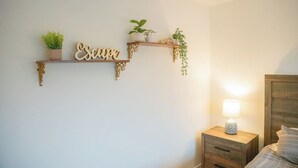 A bedroom with a bed, a nightstand with a lamp, and two wall-mounted shelves with plants and decorative items. - Guest Favorite! Easy Walk to Beach - Food & Shops! (Oceanside)