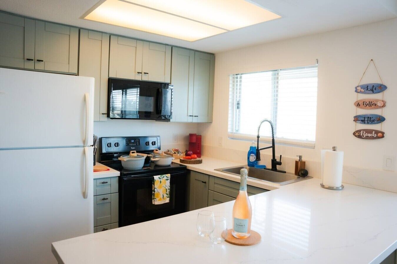 A modern kitchen with white countertops, a black stove, and a microwave. There is a window with blinds, a sink, and a decorative sign on the wall.