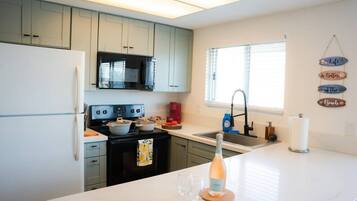A modern kitchen with white countertops, a black stove, and a microwave. There is a window with blinds, a sink, and a decorative sign on the wall.
