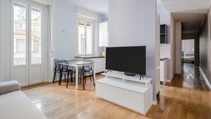 Ein modernes Wohnzimmer mit Flachbildfernseher, Essbereich mit Stühlen, einer Couch und einer Küche im Hintergrund.
