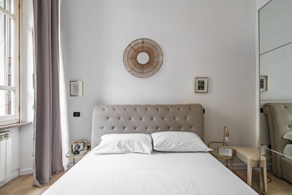 Un dormitorio con cabecera capitonada, un adorno circular en la pared, una mesita de noche con lámpara y un espejo.