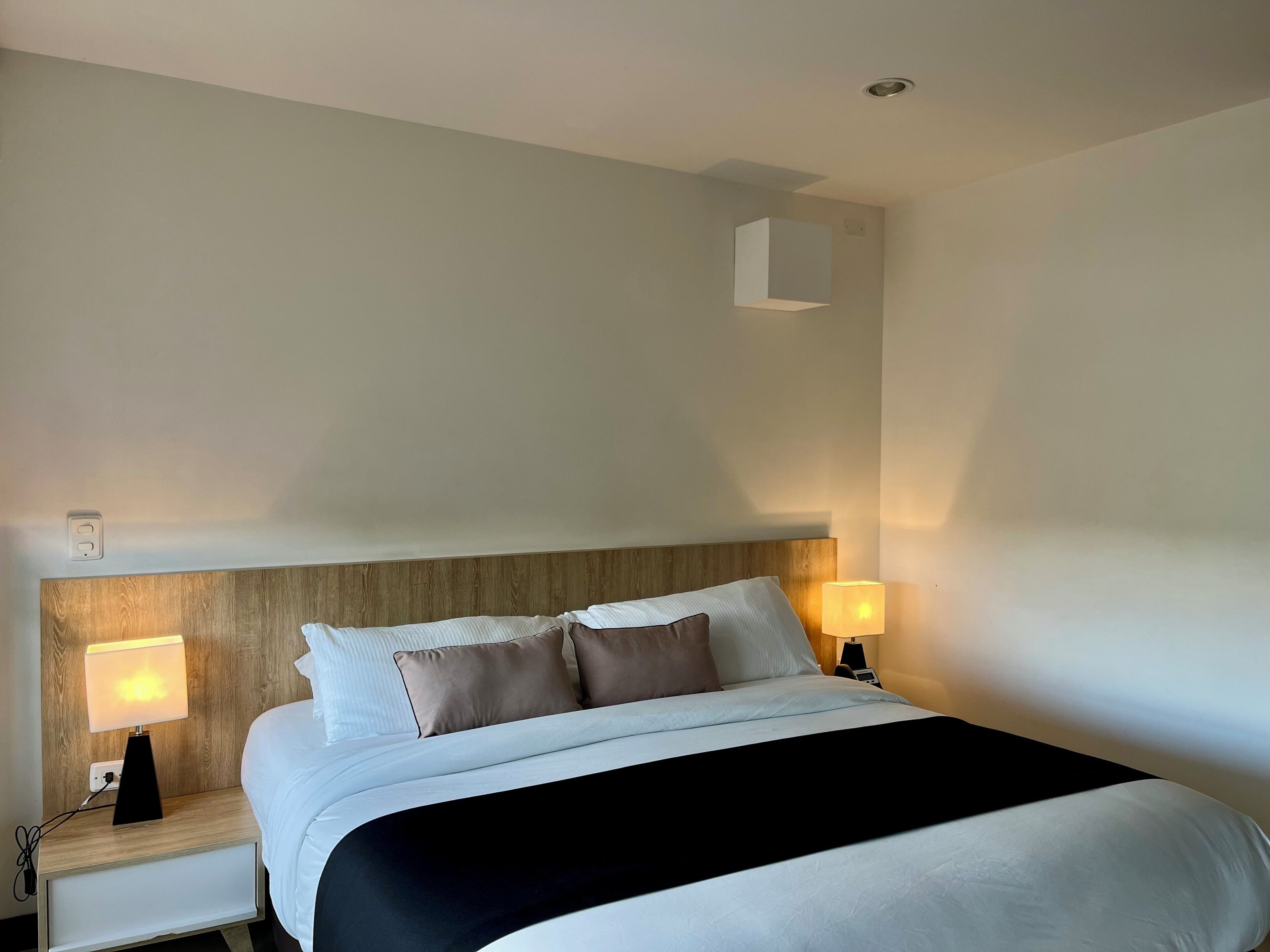 A hotel room with a wooden headboard, a bed with white bedding and a black throw, two bedside lamps, and a small wall-mounted shelf.