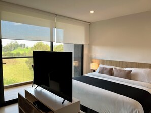 A modern hotel room with a large bed, a flat-screen TV on a wooden stand, a bedside table, and a large window offering a view of greenery.