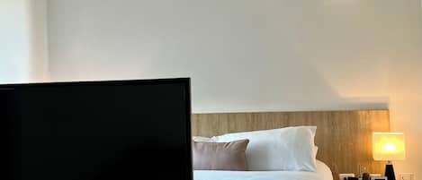 A modern hotel room with a flat-screen TV on a wooden stand, a bed with white and black bedding, a bedside table with a lamp, and a small shelf with a remote control.