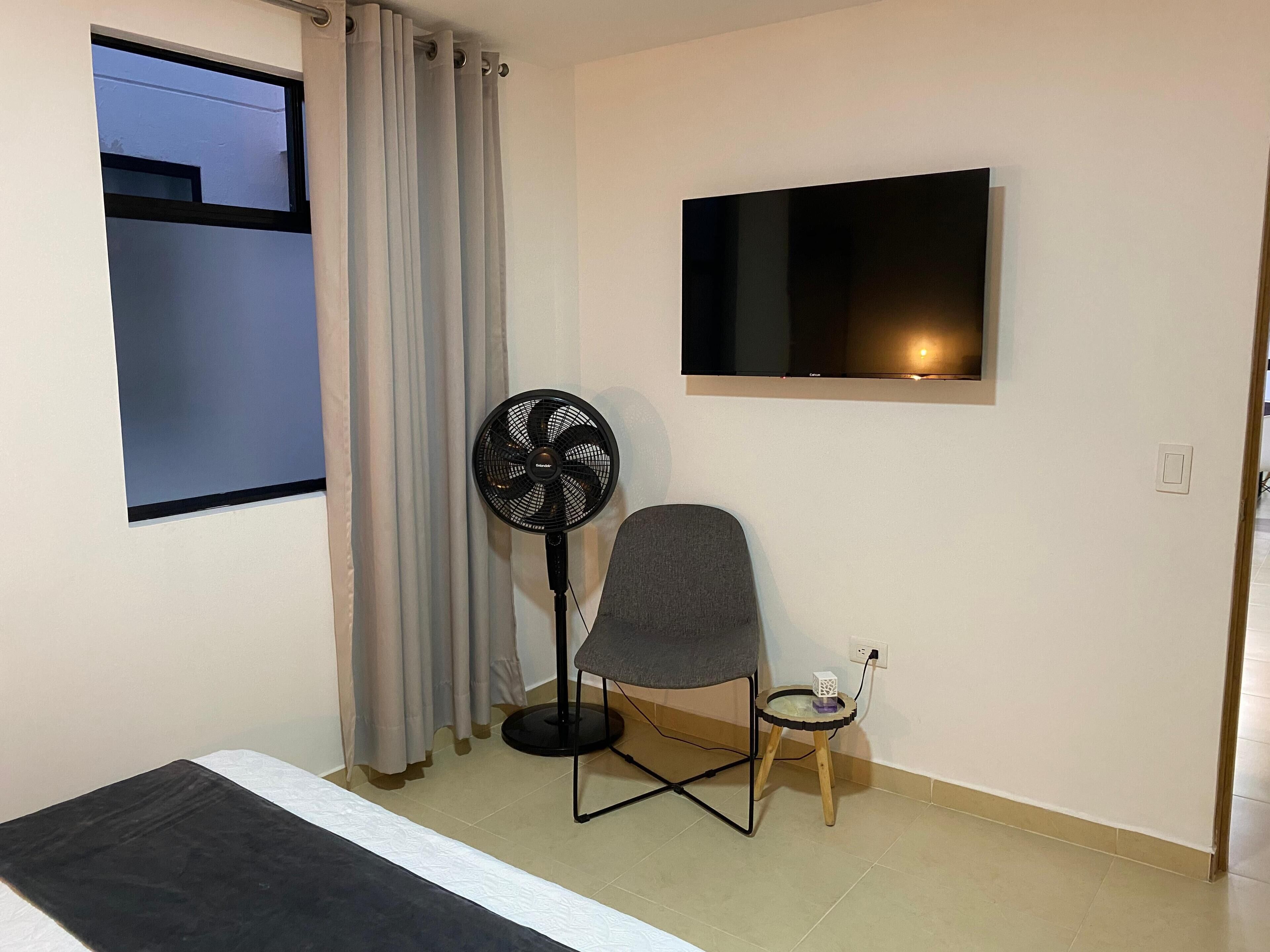 A room with a bed, a standing fan, a small table with a lamp, a TV mounted on the wall, and a window with curtains.
