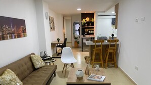 A living room with a brown sofa, white chairs, a coffee table, and a dining area with wooden chairs and a kitchen in the background. - Praia Apartaestudios (Sabaneta)