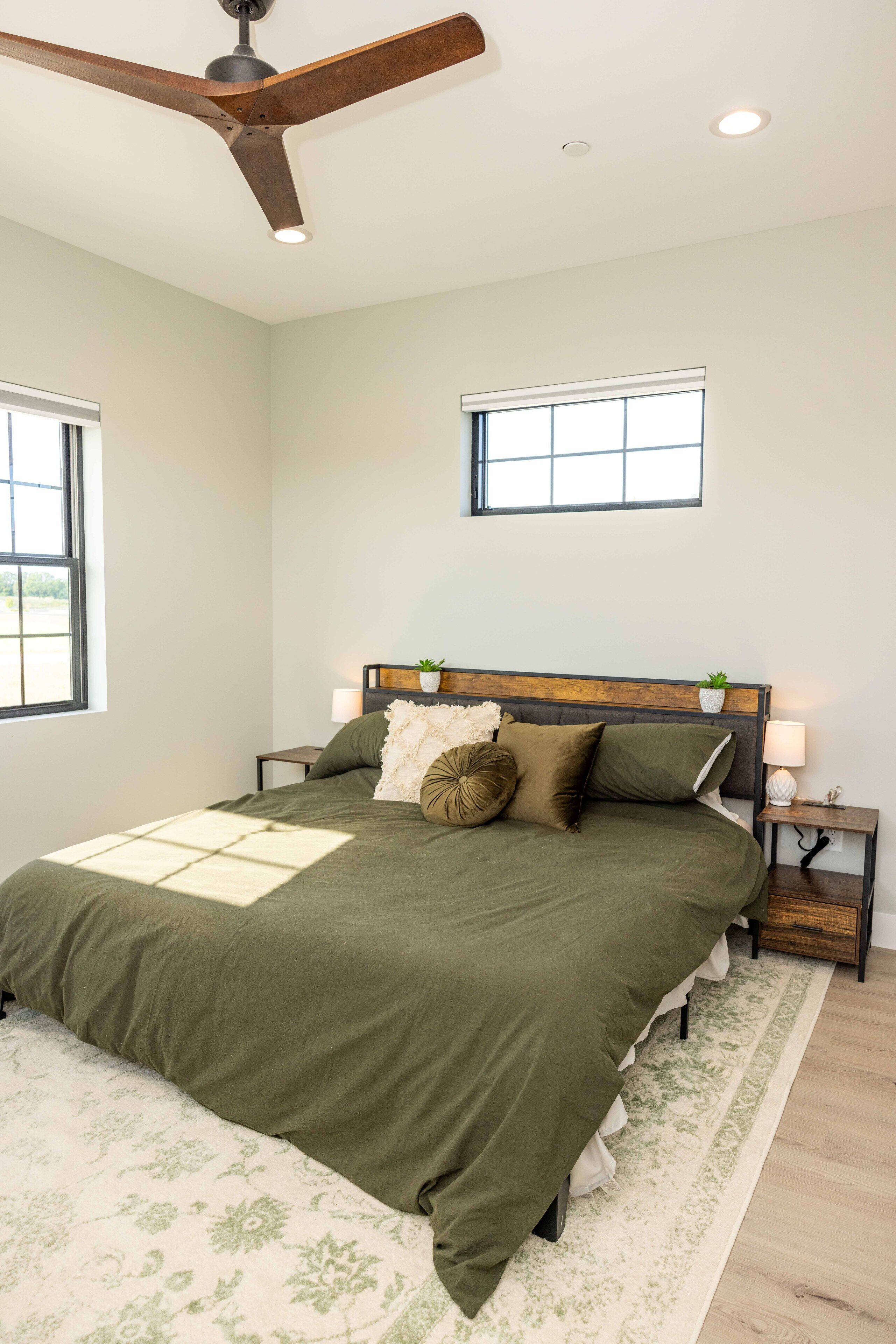 A bedroom with a bed, a ceiling fan, a nightstand, a lamp, a window, and a small plant.