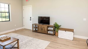 A spacious room with a wooden floor, a large window, a flat-screen TV on a wooden cabinet, and a white door. - Elderberry Farms Estate Farmstay (Traverse City)