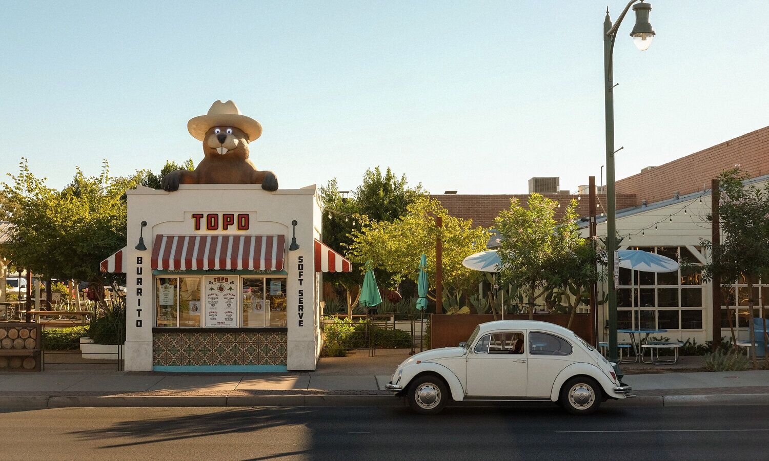 A vintage car parked in front of a small food stand named 'TOPO BURRITO' with a bear mascot and a 'SOFT SERVE' sign.