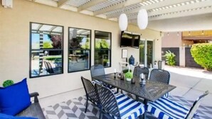 An outdoor patio area with a table and chairs, a TV mounted on the wall, and a view of the outdoors through large windows. - Stunning Heated Pool +Game Room +Great Location
 (Gilbert)