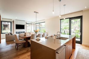 Une cuisine moderne dotée d’un grand îlot central, d’un coin repas avec des chaises, d’un téléviseur fixé au mur et de grandes fenêtres offrant une vue sur l’extérieur.