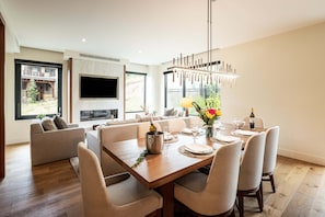 Un espace repas moderne avec une table en bois prévue pour quatre personnes, une cheminée et un grand téléviseur.