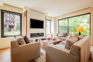 Un salon moderne doté d’un téléviseur à écran plat, de sièges confortables, d’une cheminée et de grandes fenêtres offrant une vue sur un immeuble et la verdure.