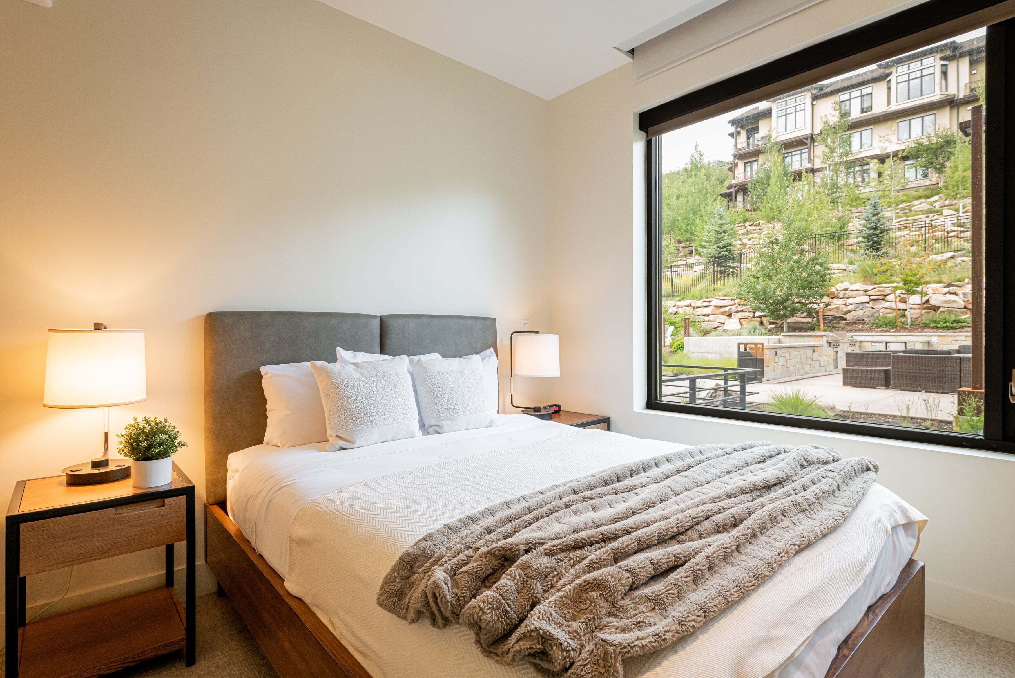 A hotel room with a large bed, two bedside tables with lamps, a view of a building and greenery outside.
