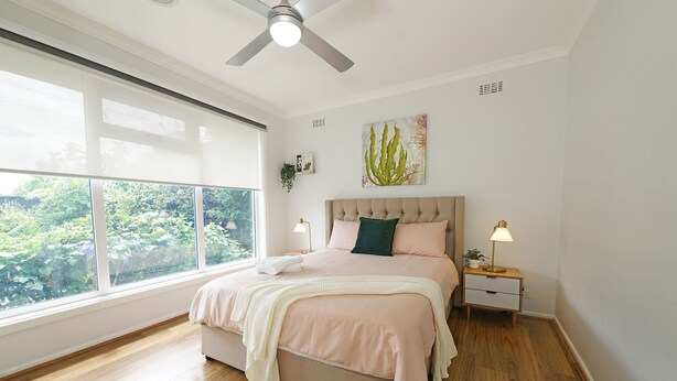 Une chambre avec une grande fenêtre, un lit doté d’une tête de lit beige, un ventilateur de plafond et une table de chevet avec une lampe.
