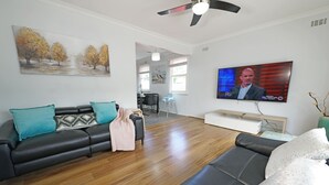 Un salon moderne avec un canapé en cuir noir, des coussins de couleur turquoise et un plancher en bois. Un téléviseur à écran plat est fixé au mur et diffuse un programme d’information.