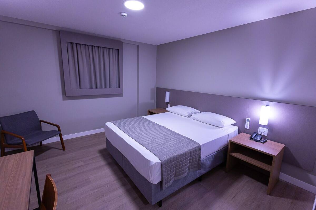 A hotel room with a bed, bedside table, chair, and a desk with a lamp.