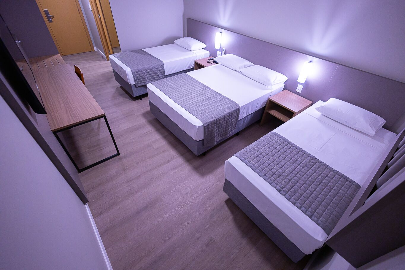 Une chambre d’hôtel avec trois lits simples, un bureau en bois et une petite table.