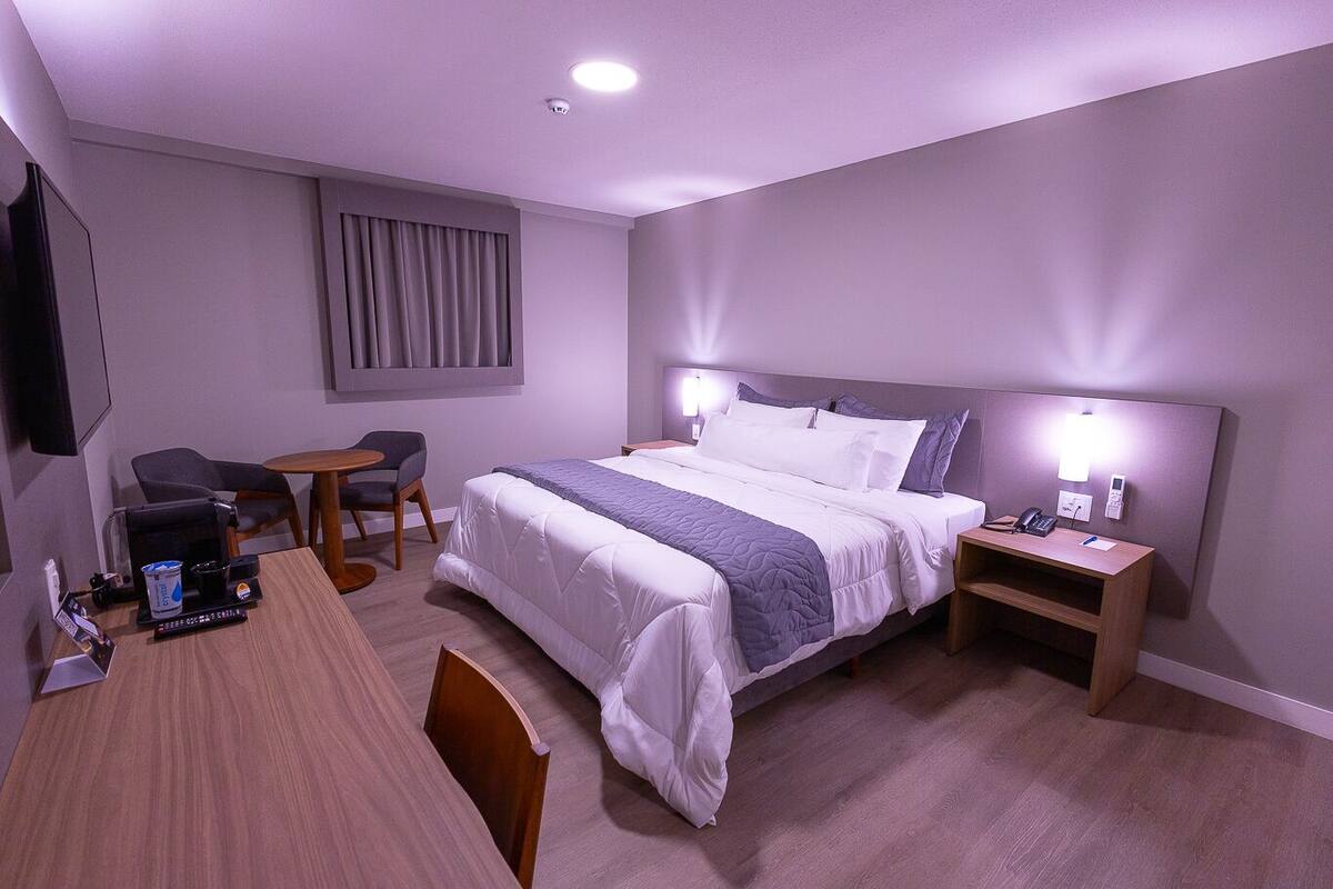 A hotel room with a bed, bedside tables, a desk with a chair, a small table, and a TV.