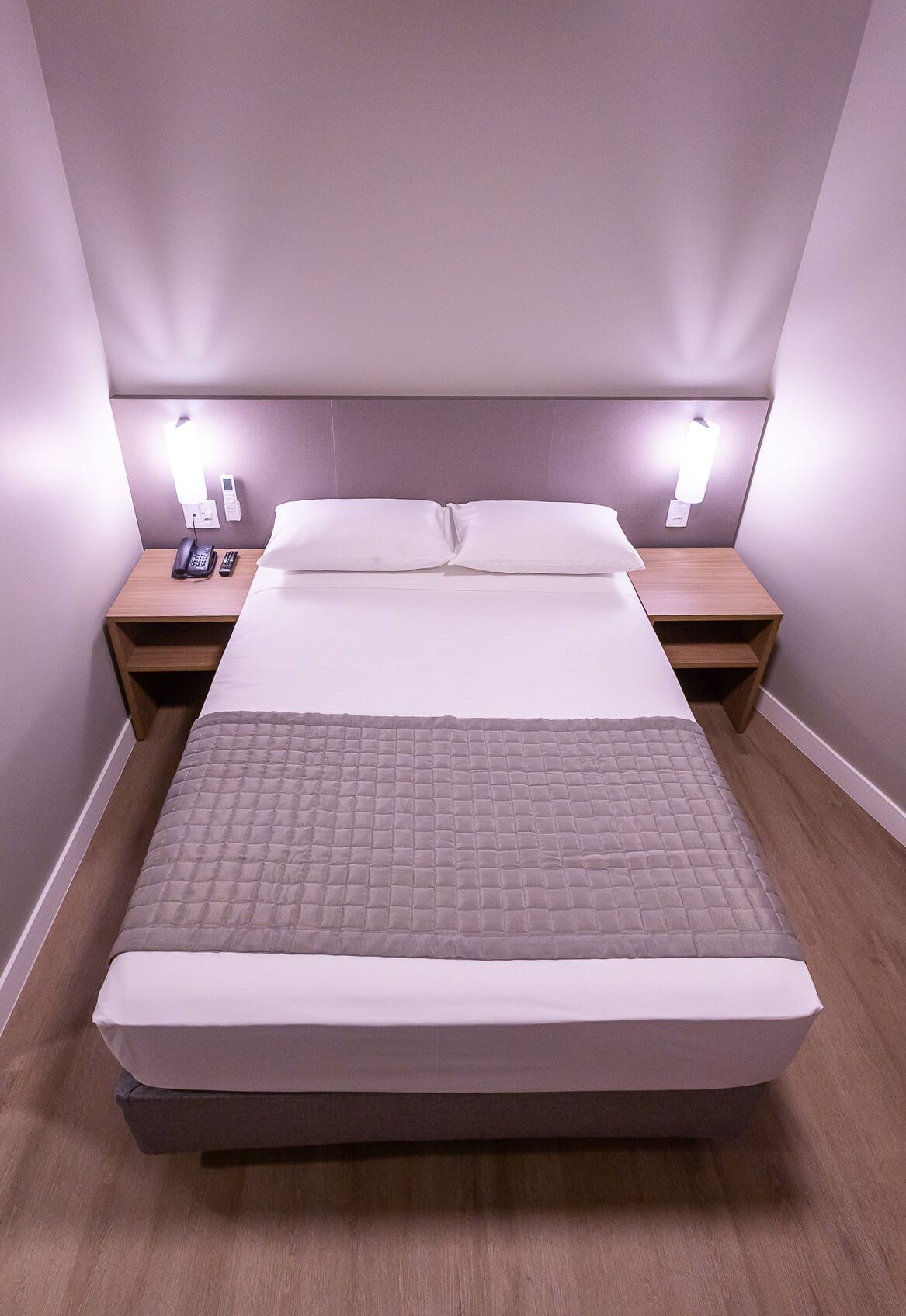 Ein ordentlich bezogenes Bett mit einer gesteppten Decke, zwei Nachttische mit Lampen und ein an der Wand befestigtes Telefon.