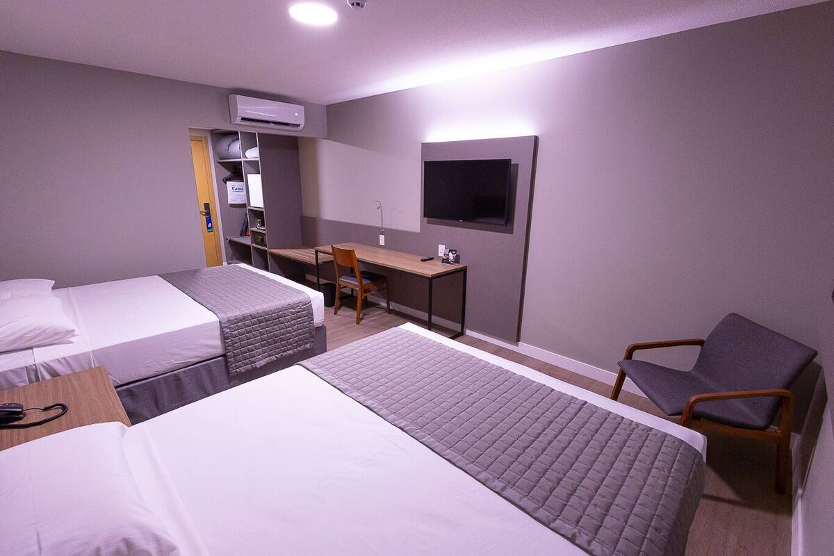 A hotel room with two beds, a desk, a chair, a TV, and an air conditioning unit.