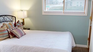 Una cama bien hecha con almohadones estampados, una lámpara de noche y una ventana con persianas.
