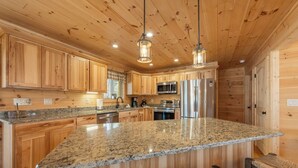 A spacious kitchen with granite countertops, wooden cabinets, stainless steel appliances, and hanging pendant lights.