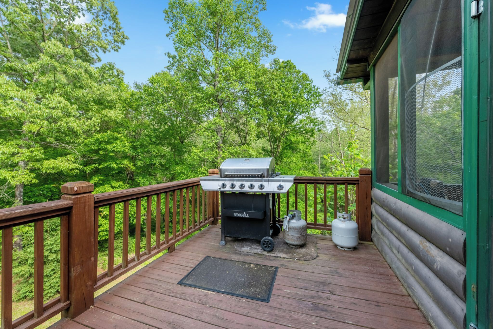 A wooden deck with a grill and propane tank, surrounded by lush greenery.