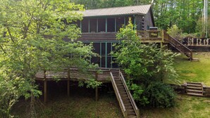 Une cabane dotée d’une terrasse et d’un escalier menant à un deuxième niveau, entourée d’une forêt dense.