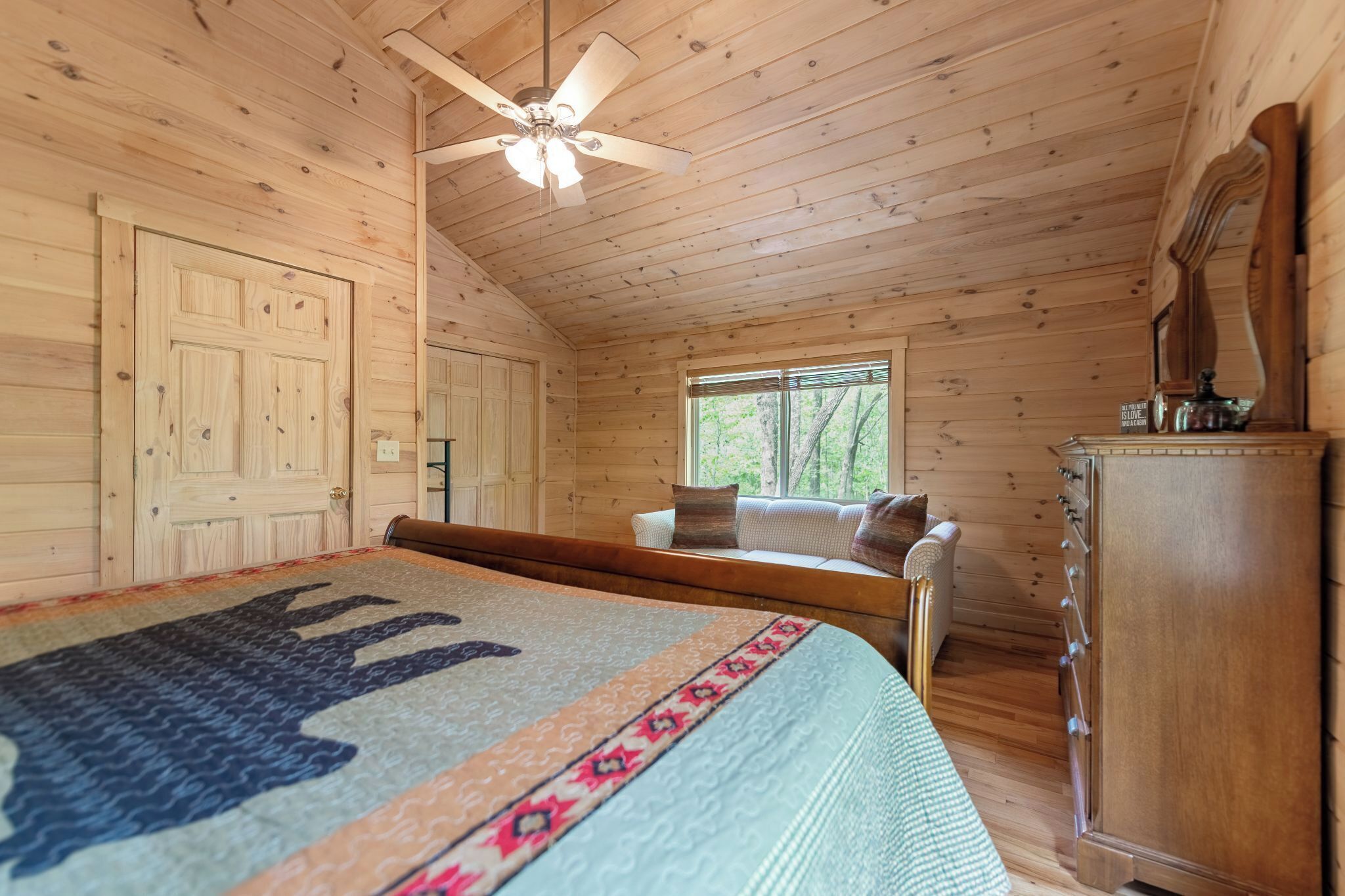 Ein Schlafzimmer in einer Holzhütte mit einem Bett, einem Fenster, einem Deckenventilator und einer Kommode.