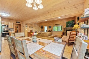 A wooden cabin interior with a dining area, kitchen, living room, and a fireplace.