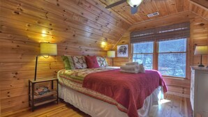 Ein Schlafzimmer einer Holzhütte mit einem Bett, Nachttischlampen, einem Fenster und einem Deckenventilator.