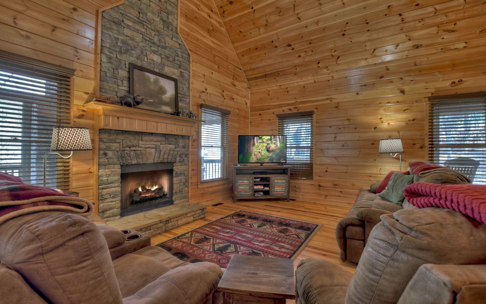 A cozy log cabin interior with a stone fireplace, a flat-screen TV, and comfortable seating.
