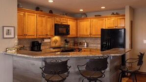 A kitchen with wooden cabinets, a black refrigerator, a microwave, and a coffee maker. There are two bar stools and a small island with a sink. - Eolus Ski-in/Ski-Out Condo w/ private hot tub/deck and Awesome Mountain Views (Durango)